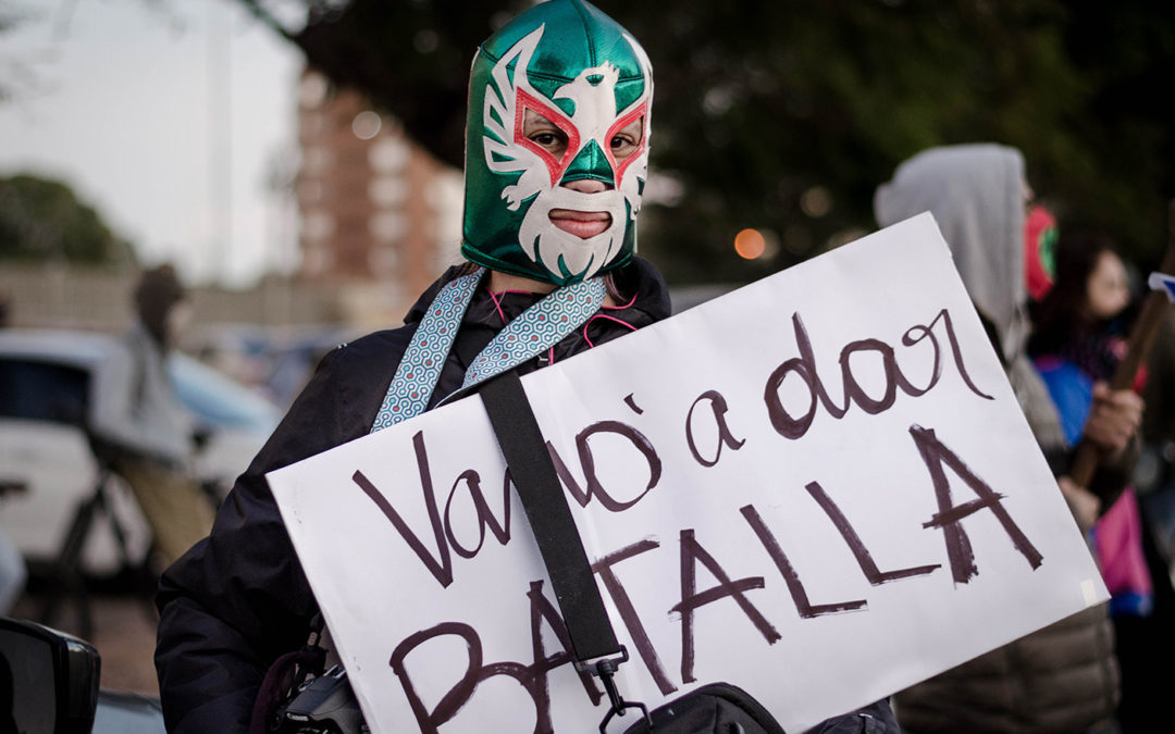 Fotógrafa manifestante en movilización en contra de la LUC. Sostiene un cartel que dice: Vamo a dar batalla y lleva puesta una máscara similar a las de lucha libre.