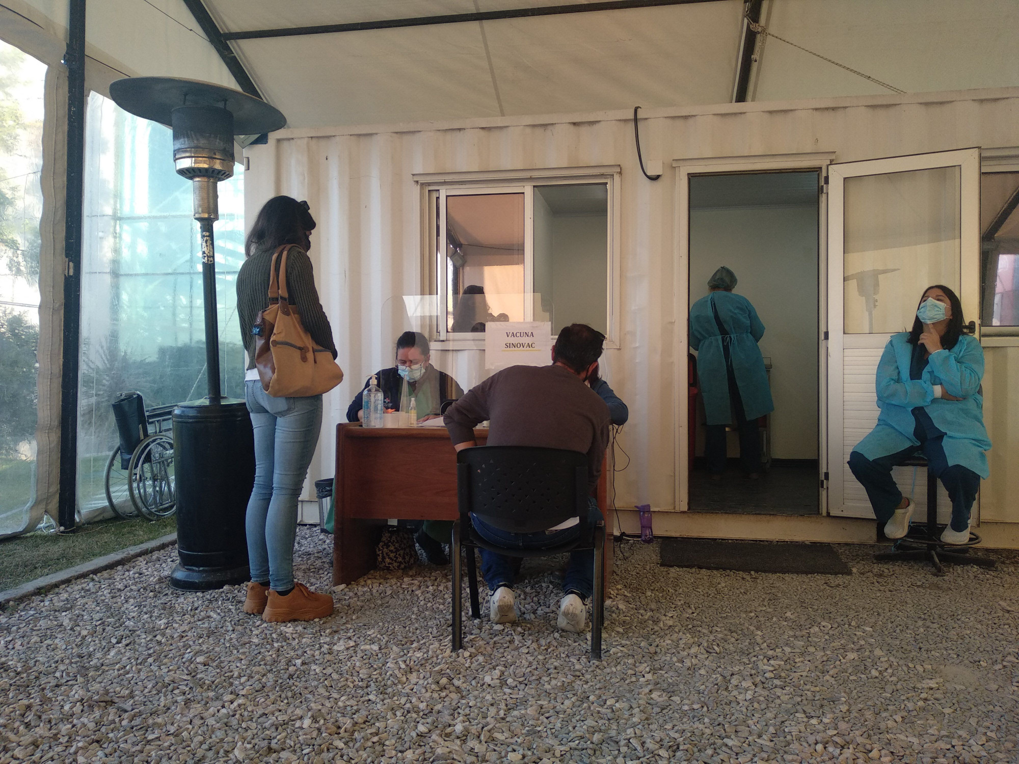 Una mujer de pie y un hombre sentado esperando para la vacunación. Tres funcionarias trabajando.
